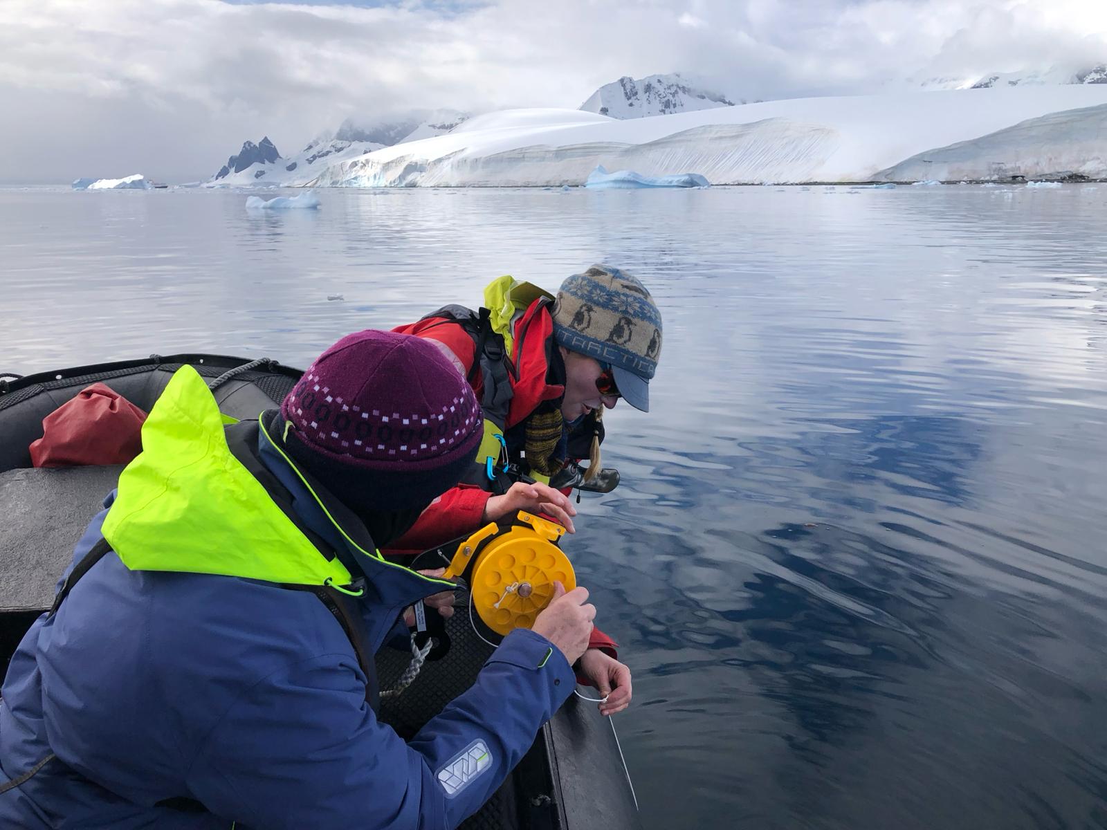 Travelers deploying a yellow Niskin bottle to collect a water sample from a calm Antarctic bay