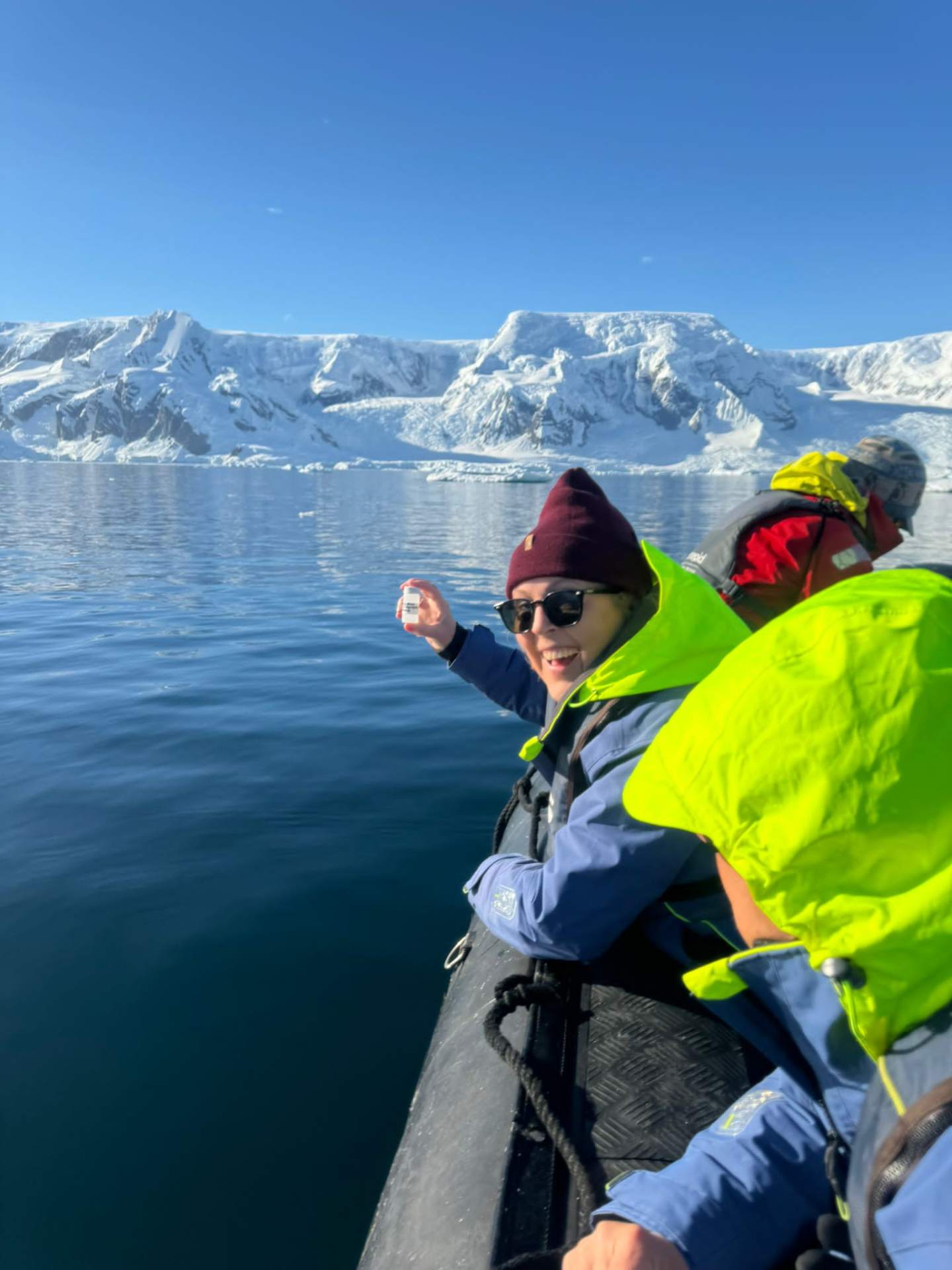 Smiling traveler holding a plankton sample vial on a sunlit Zodiac with snowy Antarctic mountains