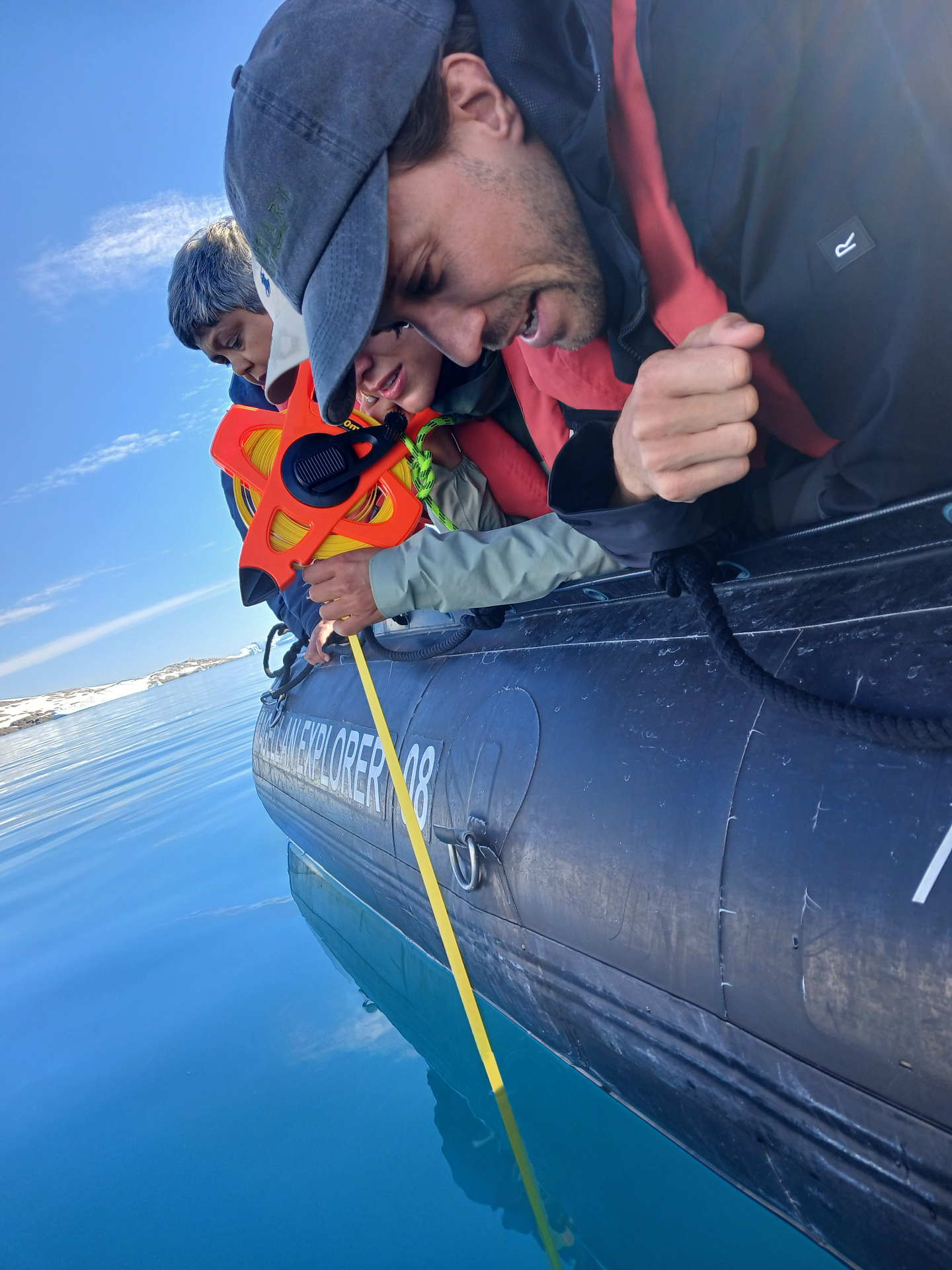 Two travelers leaning over a Zodiac to deploy an orange Secchi disk reel into mirror-calm Antarctic water