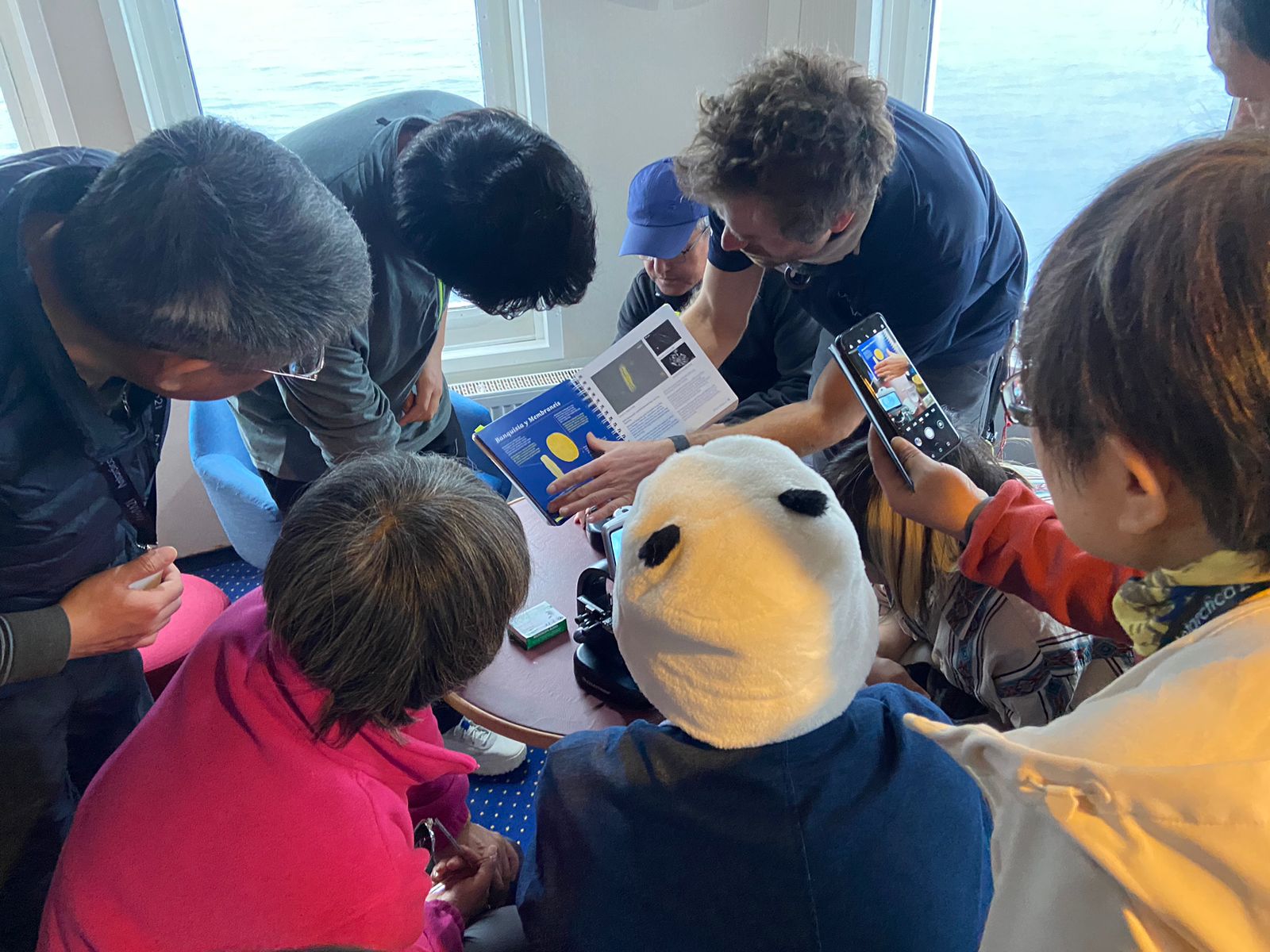 Onboard workshop where travelers gather around a plankton field guide and microscope demonstration