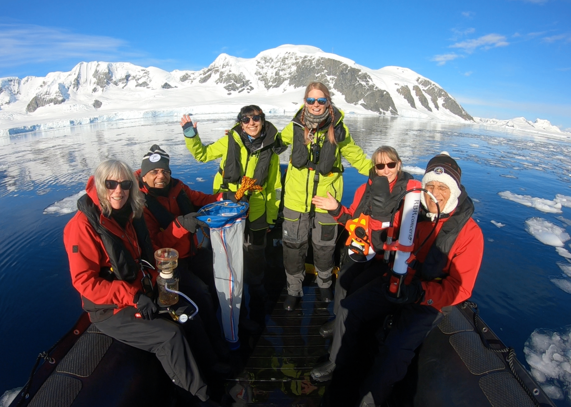 Group of researchers on a Zodiac boat in front of snowy Antarctic mountains with sampling equipment